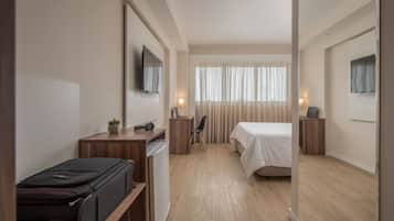 Quarto de hotel com cama, escrivaninha, cadeira e mala.