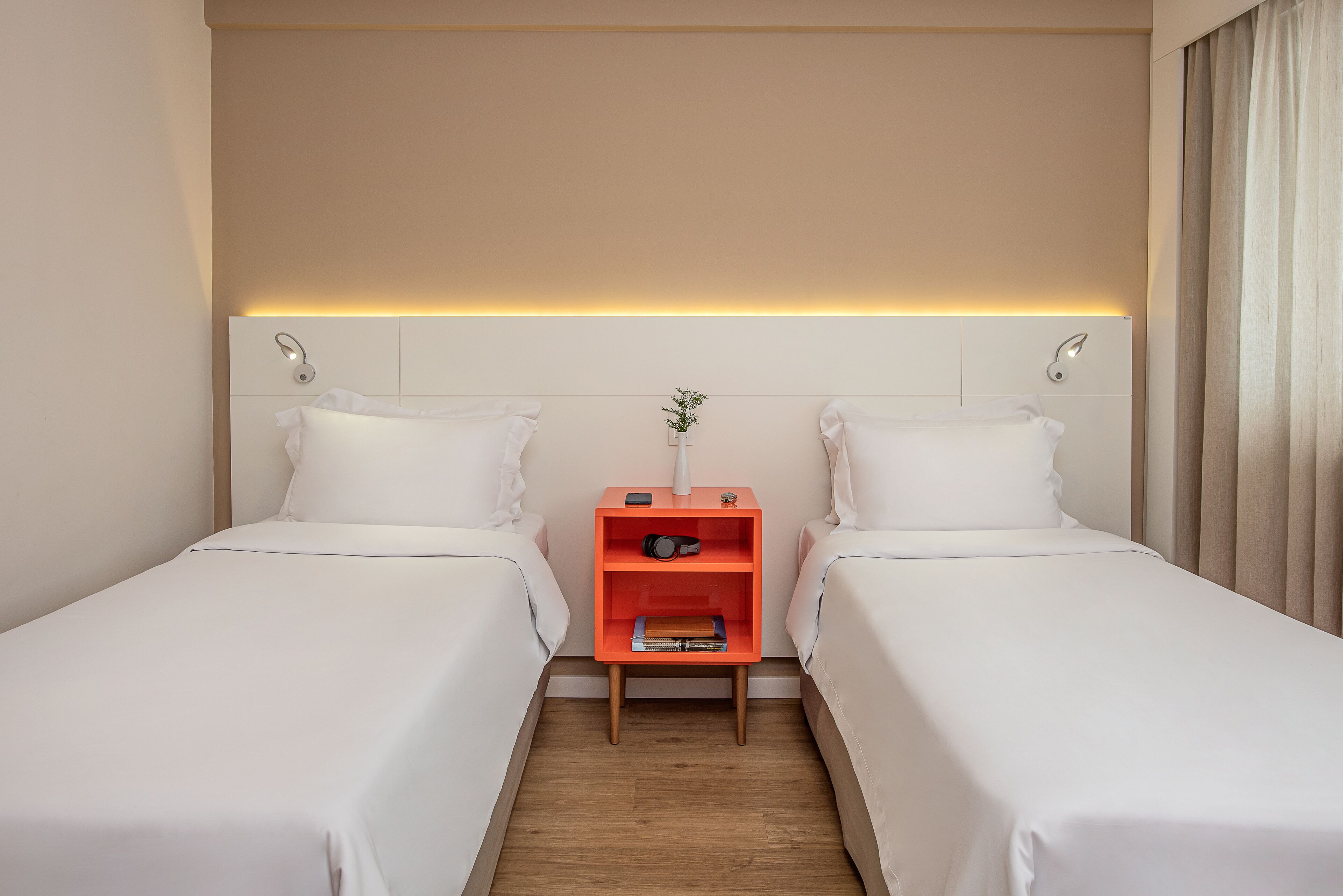 Una habitación de hotel con dos camas individuales, una mesita de noche naranja pequeña con una planta y libros, y una mesita de noche con una lámpara.
