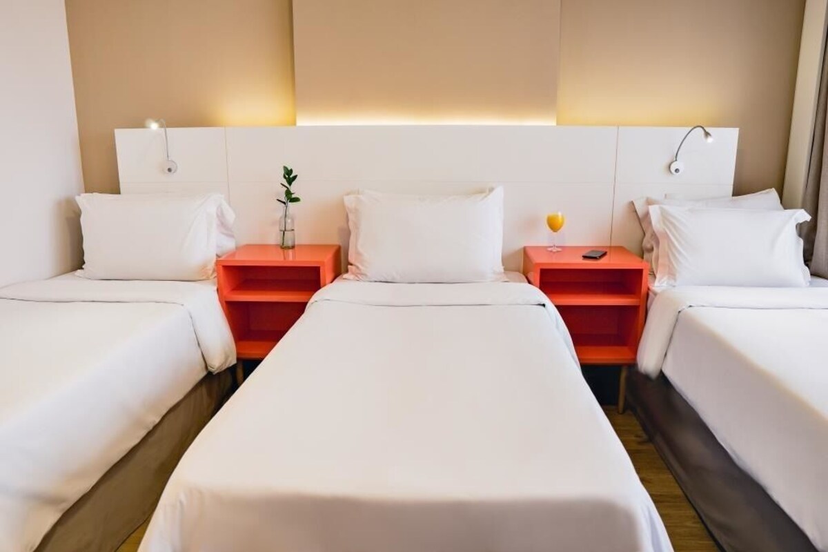 Habitación de hotel con dos camas individuales, cada una con colchón y almohada blancos, y mesitas de noche naranjas a ambos lados.