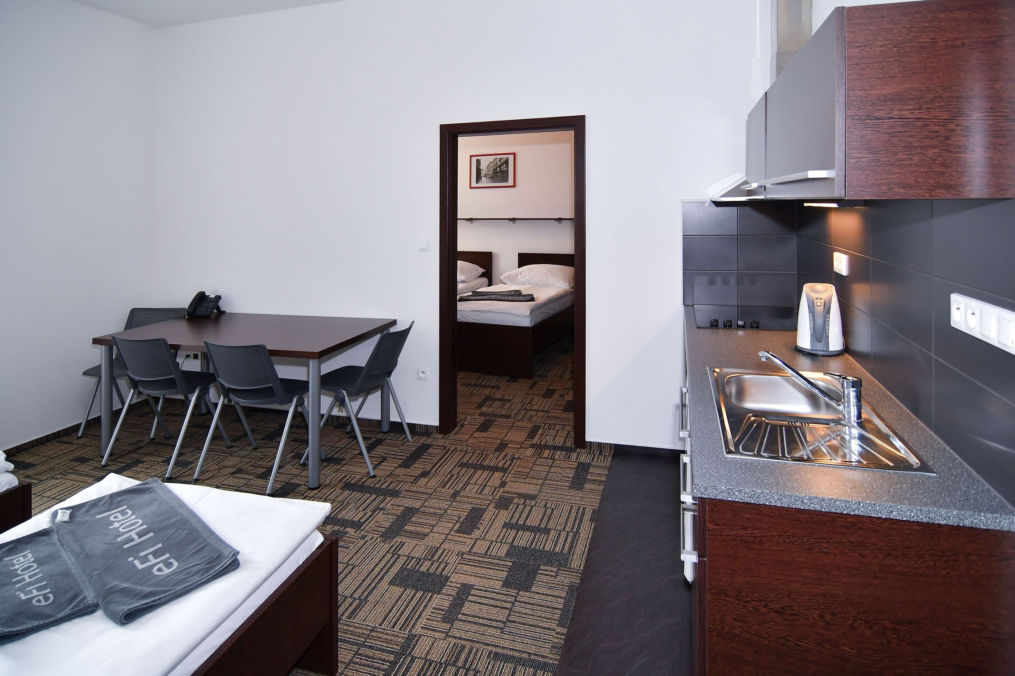 Habitación de hotel pequeña con cama, kitchenette, zona de comedor con sillas y baño visible a través de una puerta abierta.