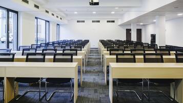 Une salle de conférence avec des rangées de tables et de chaises, de grandes fenêtres et un projecteur fixé au plafond.