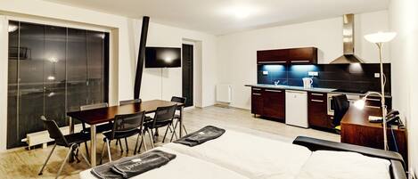 A modern hotel room with a bed, a dining table with chairs, a kitchen area, and a TV.