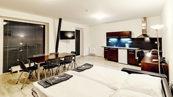 A modern hotel room with a bed, a dining table with chairs, a kitchen area, and a TV.