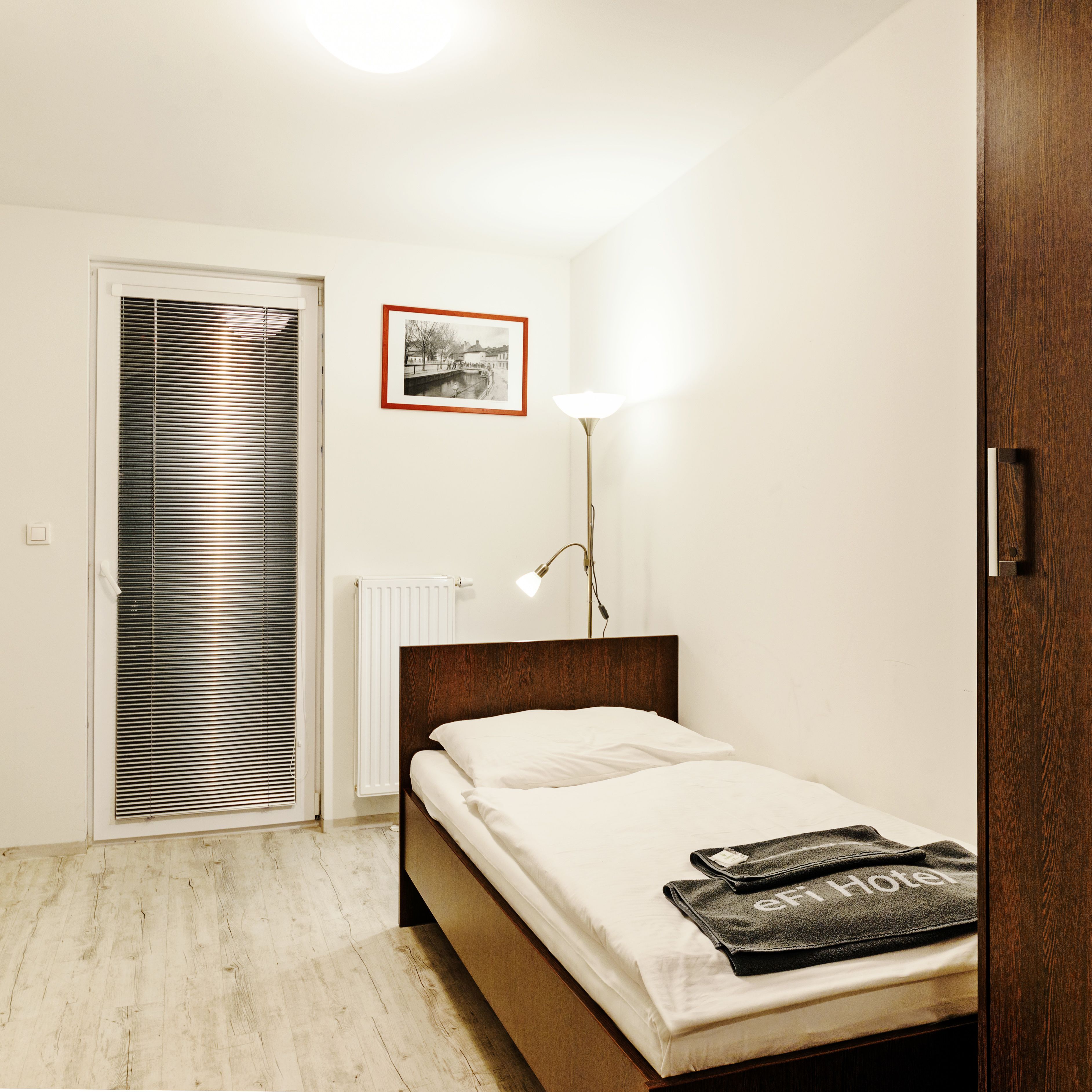 A bedroom with a bed, a radiator, a framed picture on the wall, and a floor lamp.