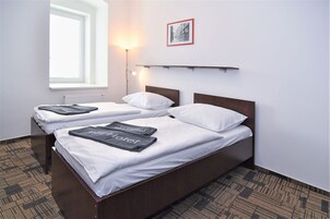 Quarto de hotel com duas camas, um radiador, uma janela, uma prateleira fixada na parede e um quadro emoldurado.