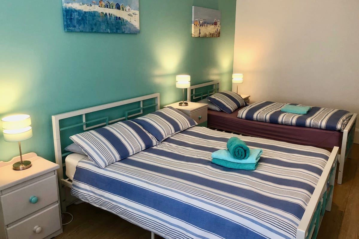 A room with two beds, each with striped bedding and a teal towel. There are bedside tables with lamps, a nightstand, and a wall-mounted painting.