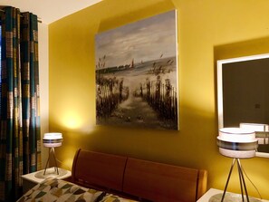 A room with a yellow wall, a painting of a beach scene, a bed with a patterned headboard, and two floor lamps.