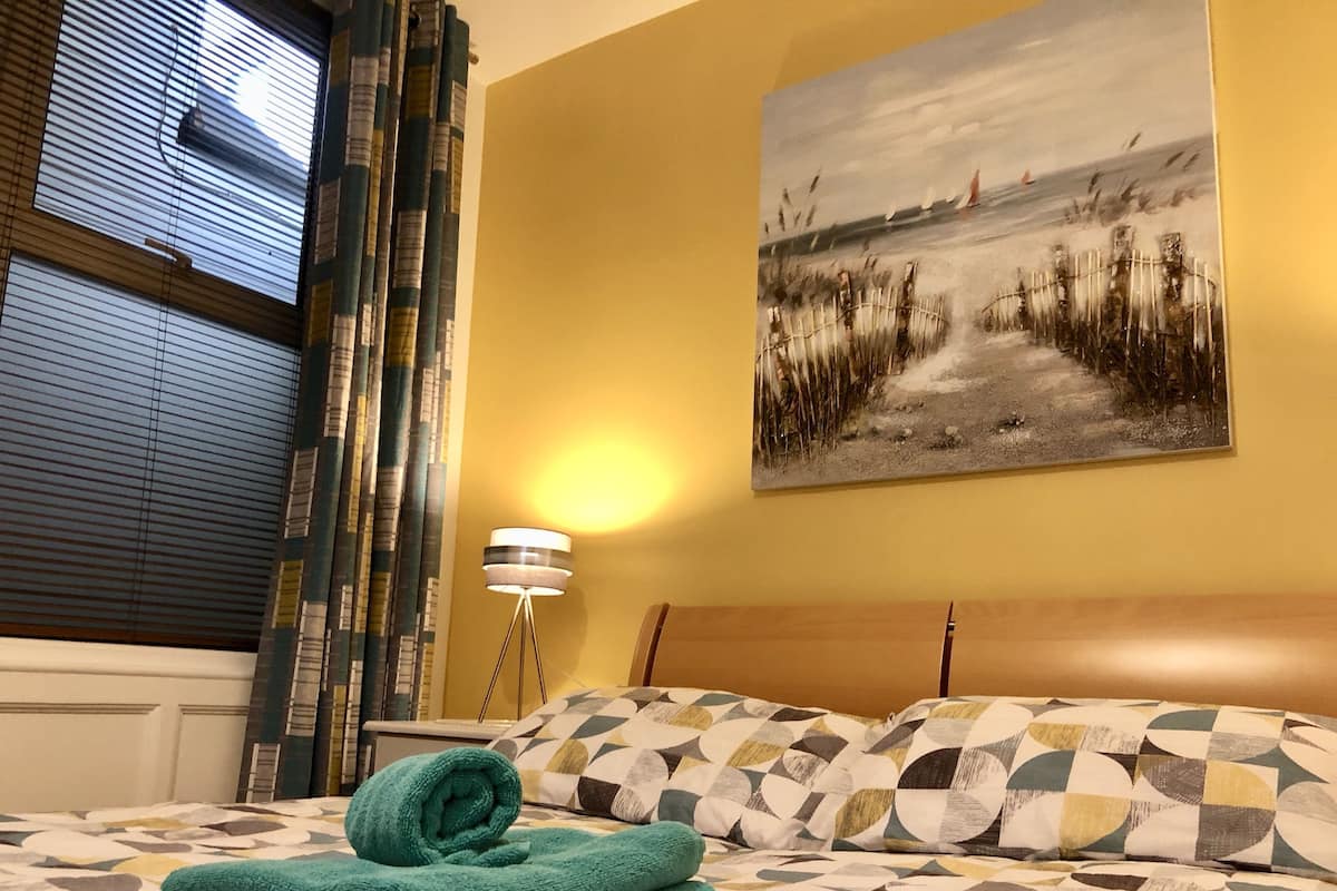 A hotel room with a bed, a painting of a landscape, a lamp, and curtains.