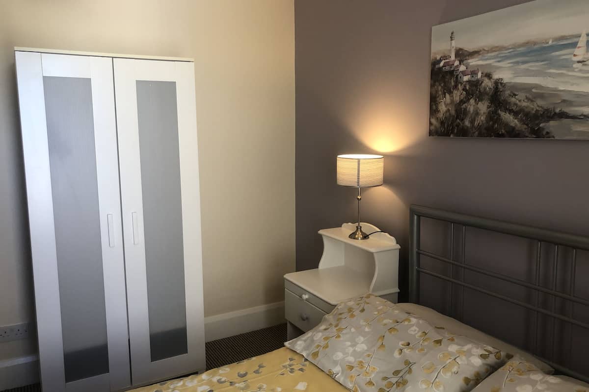 A bedroom with a double bed, a nightstand, a lamp, a wardrobe, and a painting on the wall.