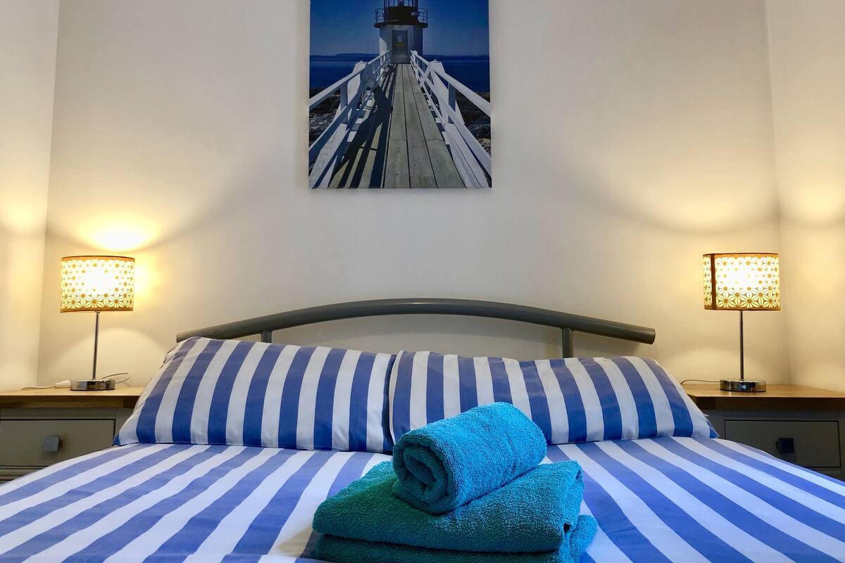 A hotel room with a bed featuring blue and white striped bedding, two bedside lamps, a painting of a lighthouse on the wall, and a neatly folded blue towel on the bed.