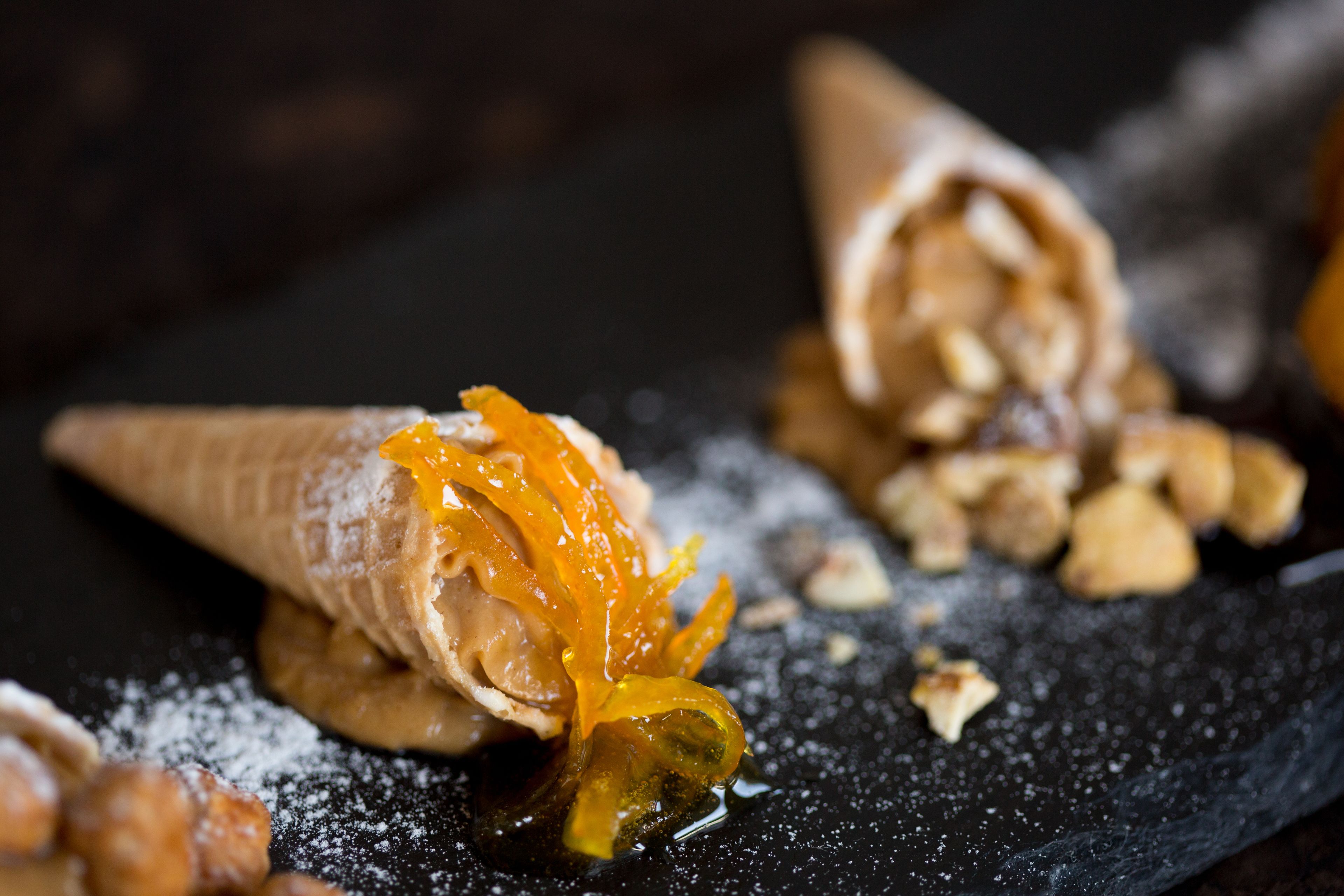 Un postre con cucurucho de gofre, ralladura de naranja y nueces sobre un plato de pizarra oscura.