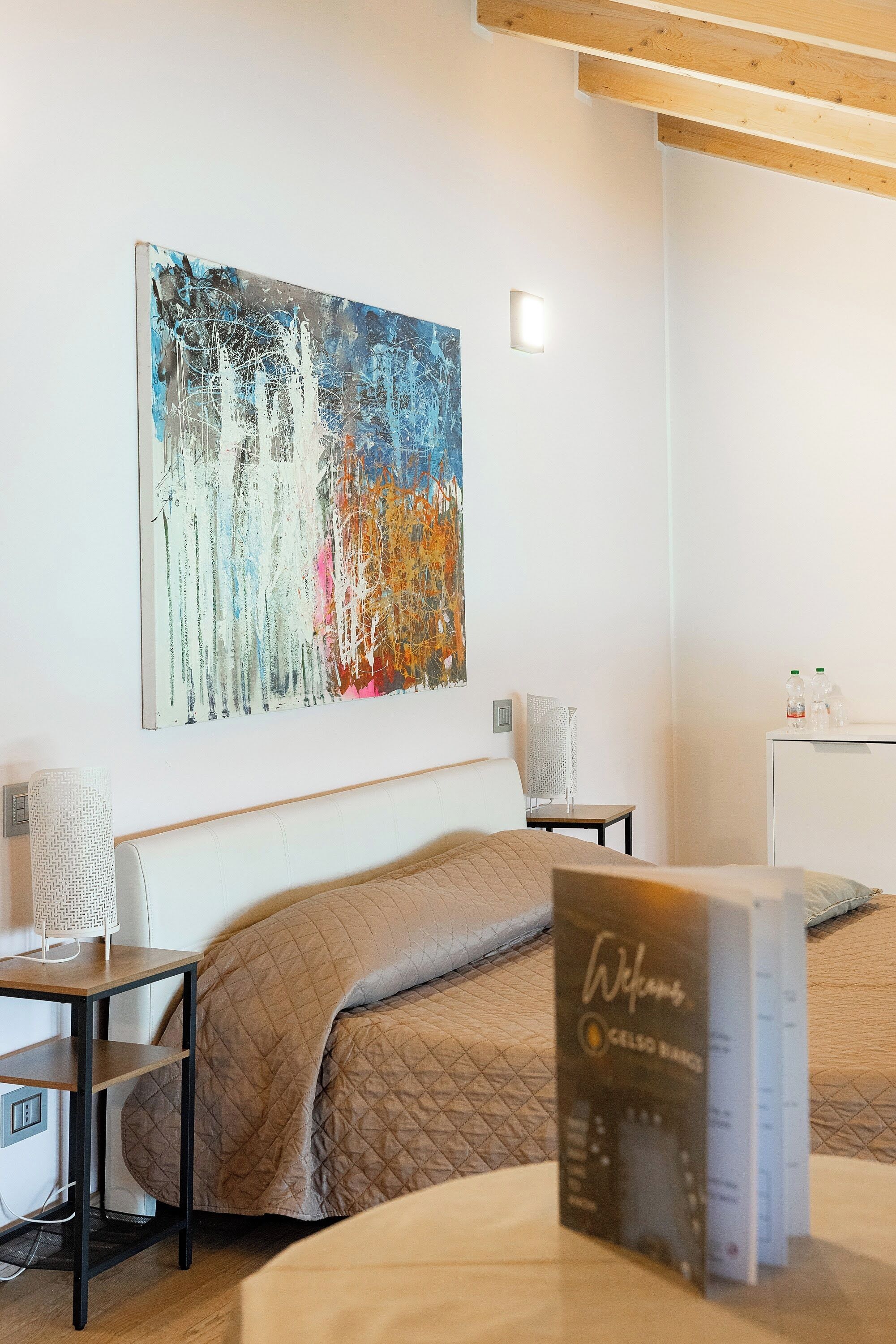 Habitación de hotel con una cama, mesita de noche, lámpara, un cuadro en la pared y un mueble pequeño con botellas.