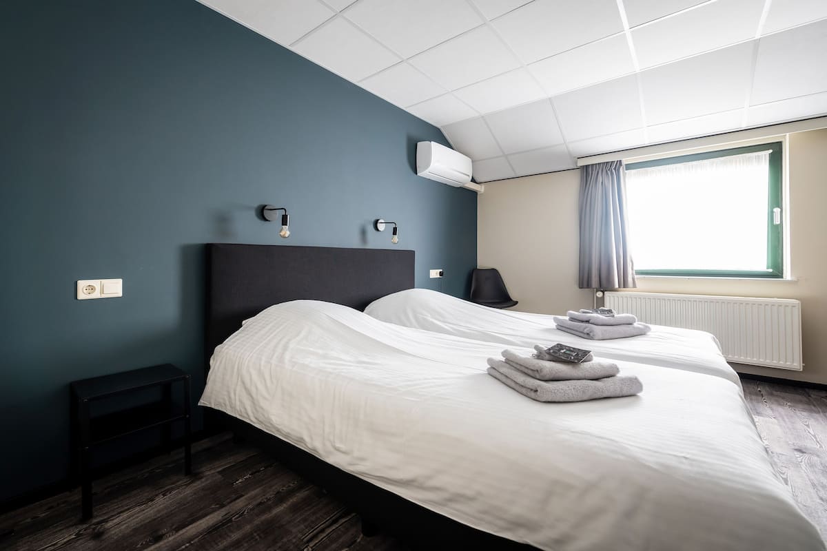 Quarto de hotel com duas camas, mesa de cabeceira, luminária, ar condicionado, janela com cortinas e radiador.