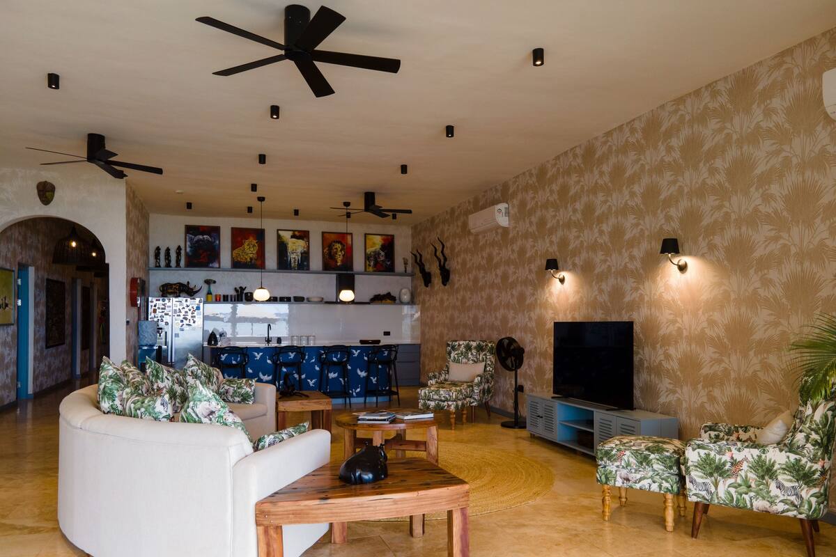 A spacious living area with a curved sofa, a coffee table, and two armchairs with tropical prints. A flat-screen TV and a ceiling fan are visible. The kitchen area is in the background with a bar counter and chairs.