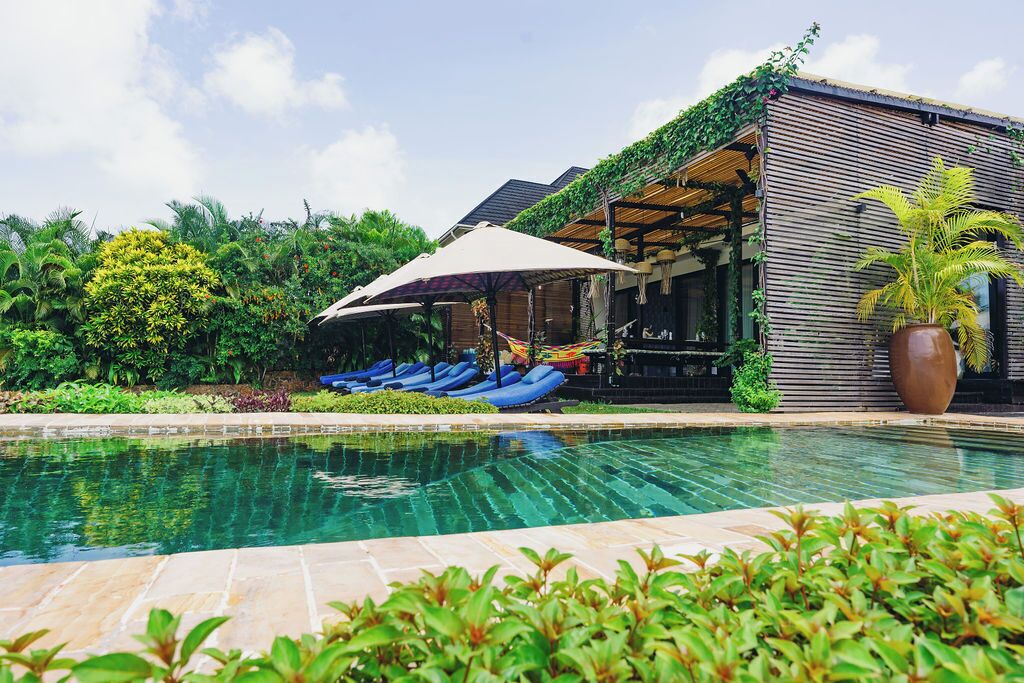 A pool area with lounge chairs and umbrellas, surrounded by lush greenery and a wooden building with a covered seating area.