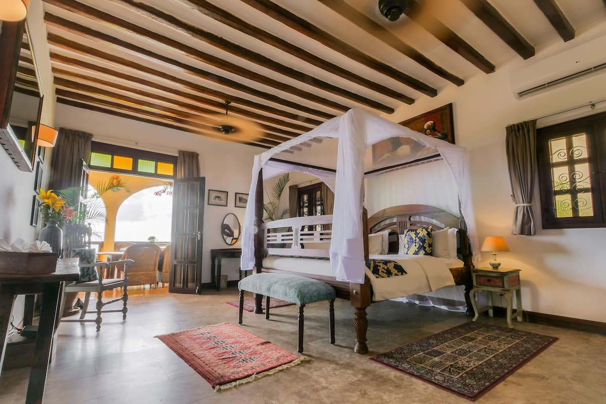 A four-poster bed with a canopy, a wooden bench, a small table with a lamp, and a view of the outdoors through a window.