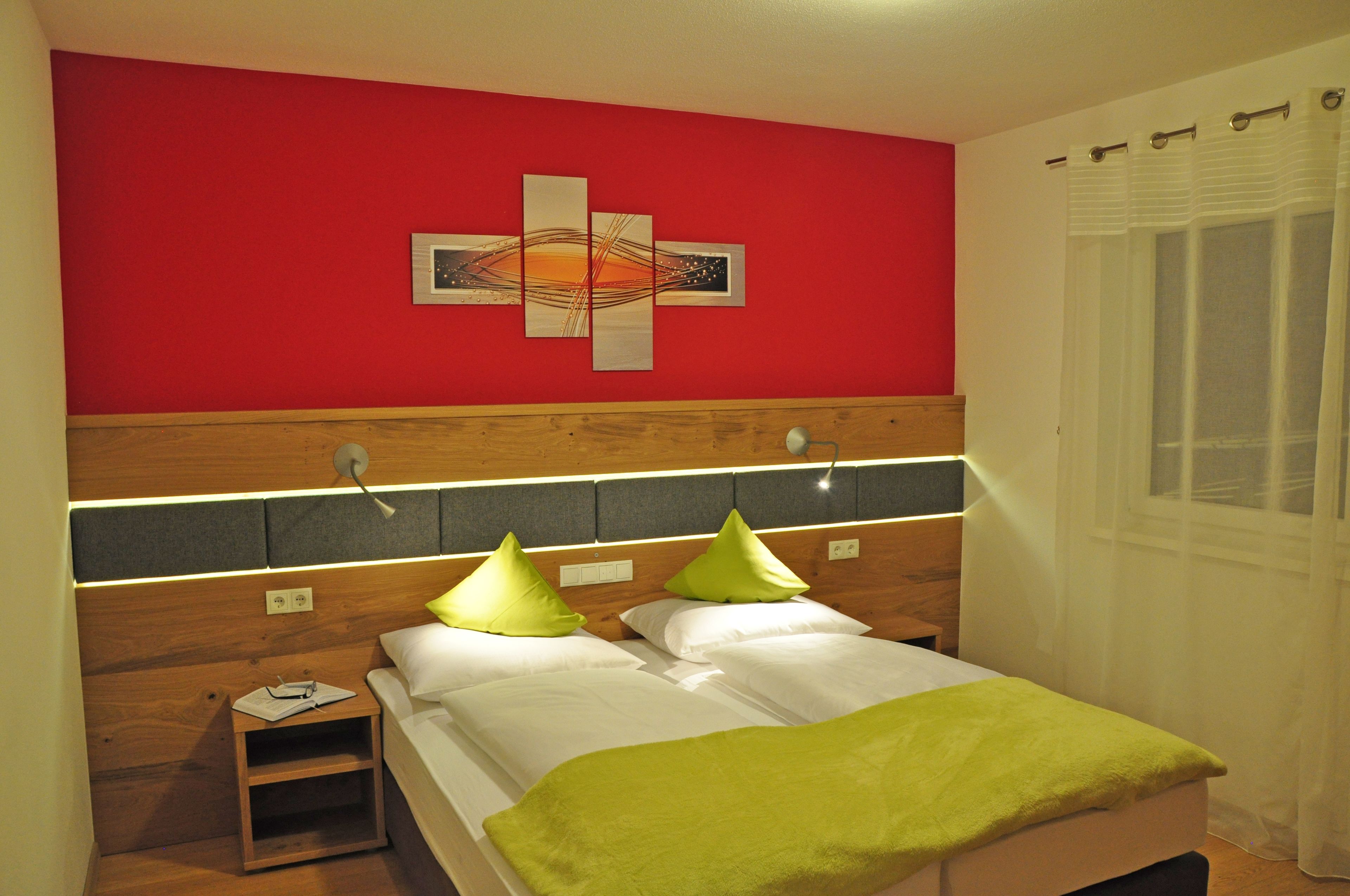 Habitación de hotel con cabecera de madera, cama con ropa blanca y almohadas verdes, mesita de noche con lámpara y pared de acento roja con una obra de arte con forma de cruz.