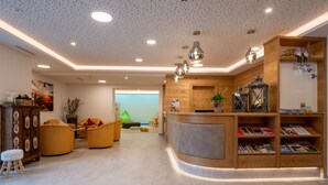 A modern reception area with a wooden reception desk, seating area with yellow chairs, and a wall-mounted shelf with magazines. - Appartement Schattauer (Wagrain)