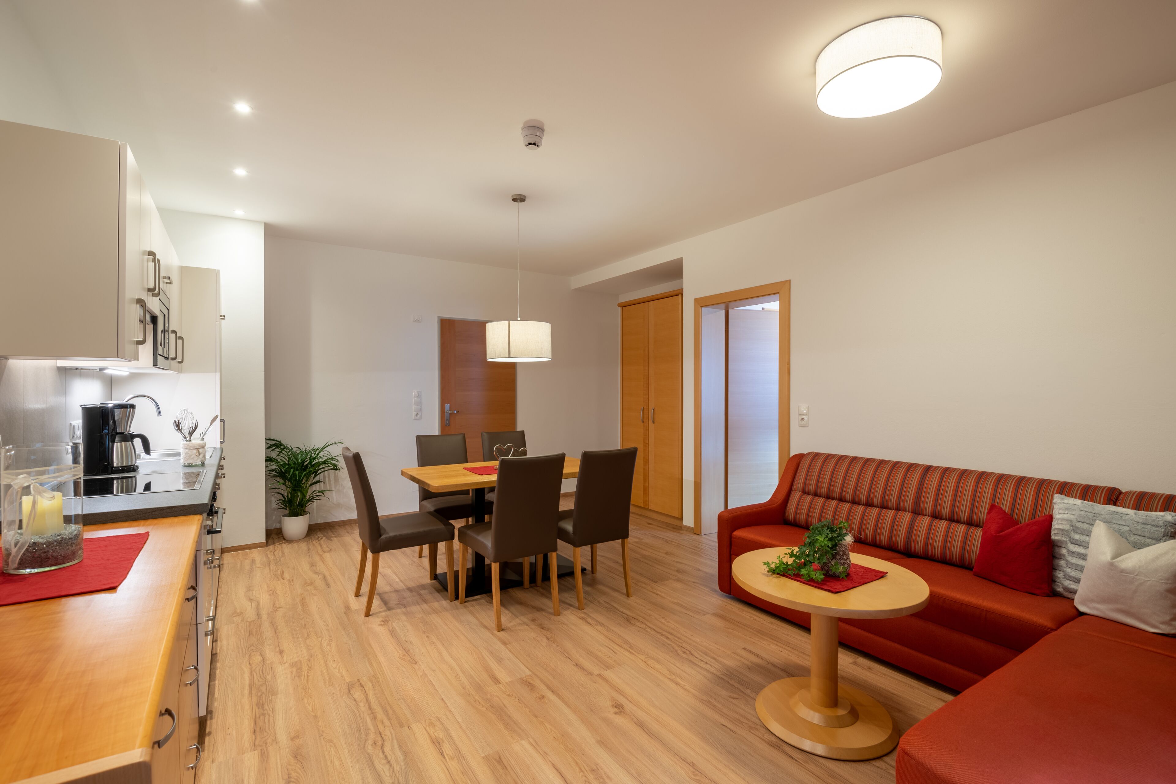 A modern living room with a sofa, a dining table with chairs, and a kitchen area with a coffee machine.