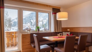 A cozy dining area with a wooden table and brown upholstered chairs, a bench, and a view of a snowy mountain landscape through large windows. - Appartement Schattauer (Wagrain)