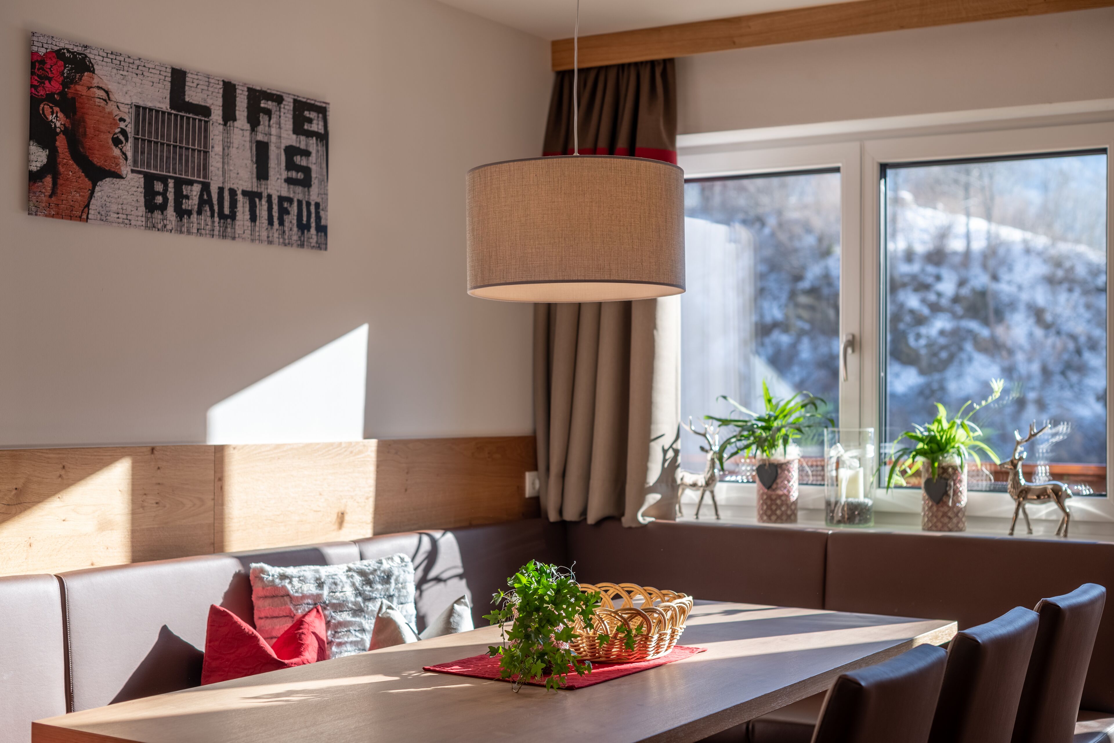 A modern dining area with a wooden table, cushioned seating, and a large window with a view of snow-covered trees.