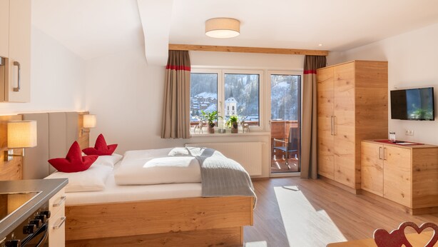 A modern hotel room with a wooden bed, a desk, a television, and a view of a snowy mountain. - Appartement Schattauer (Wagrain)