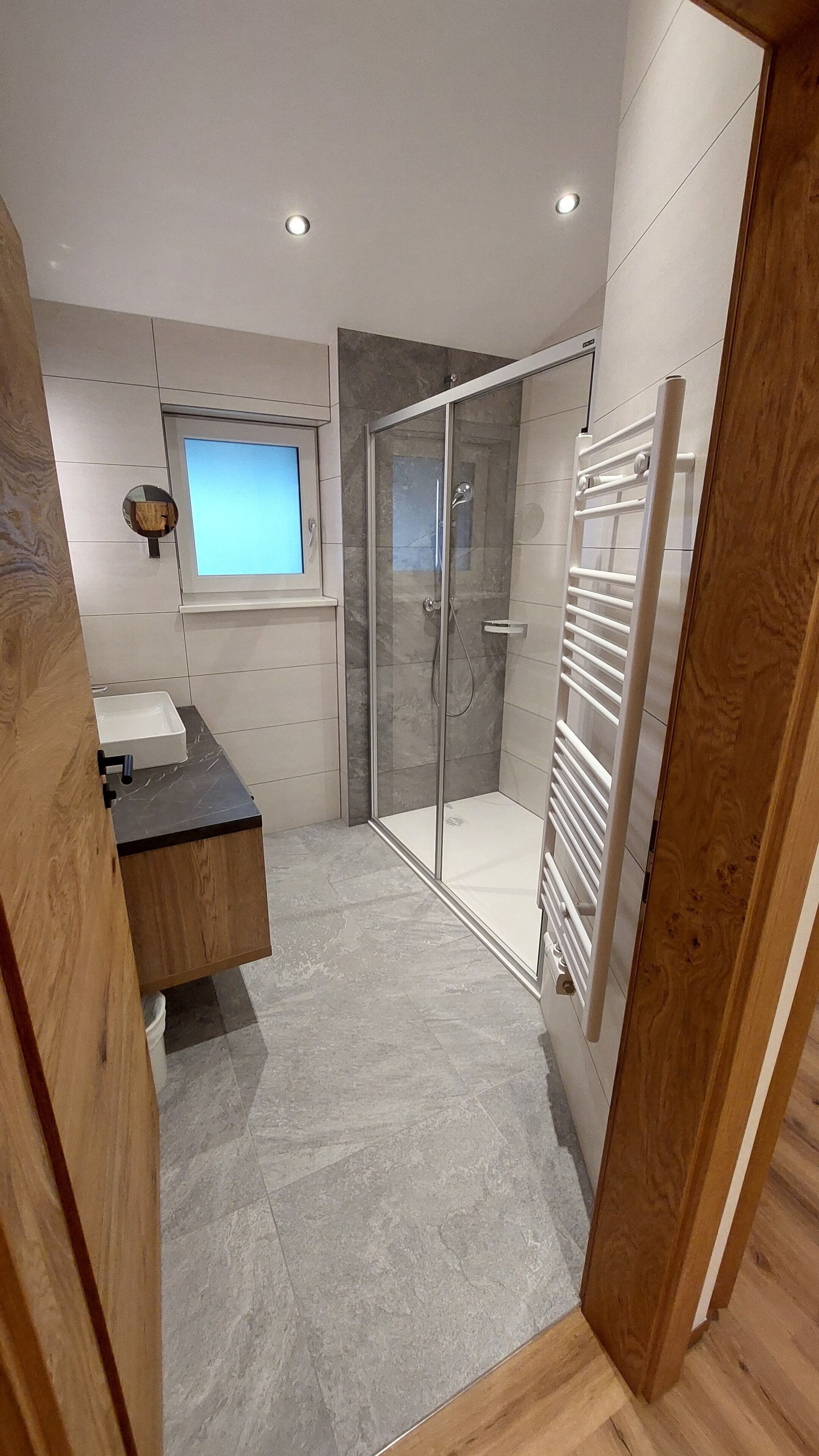 A modern bathroom with a glass shower enclosure, a wooden vanity, and a built-in window.