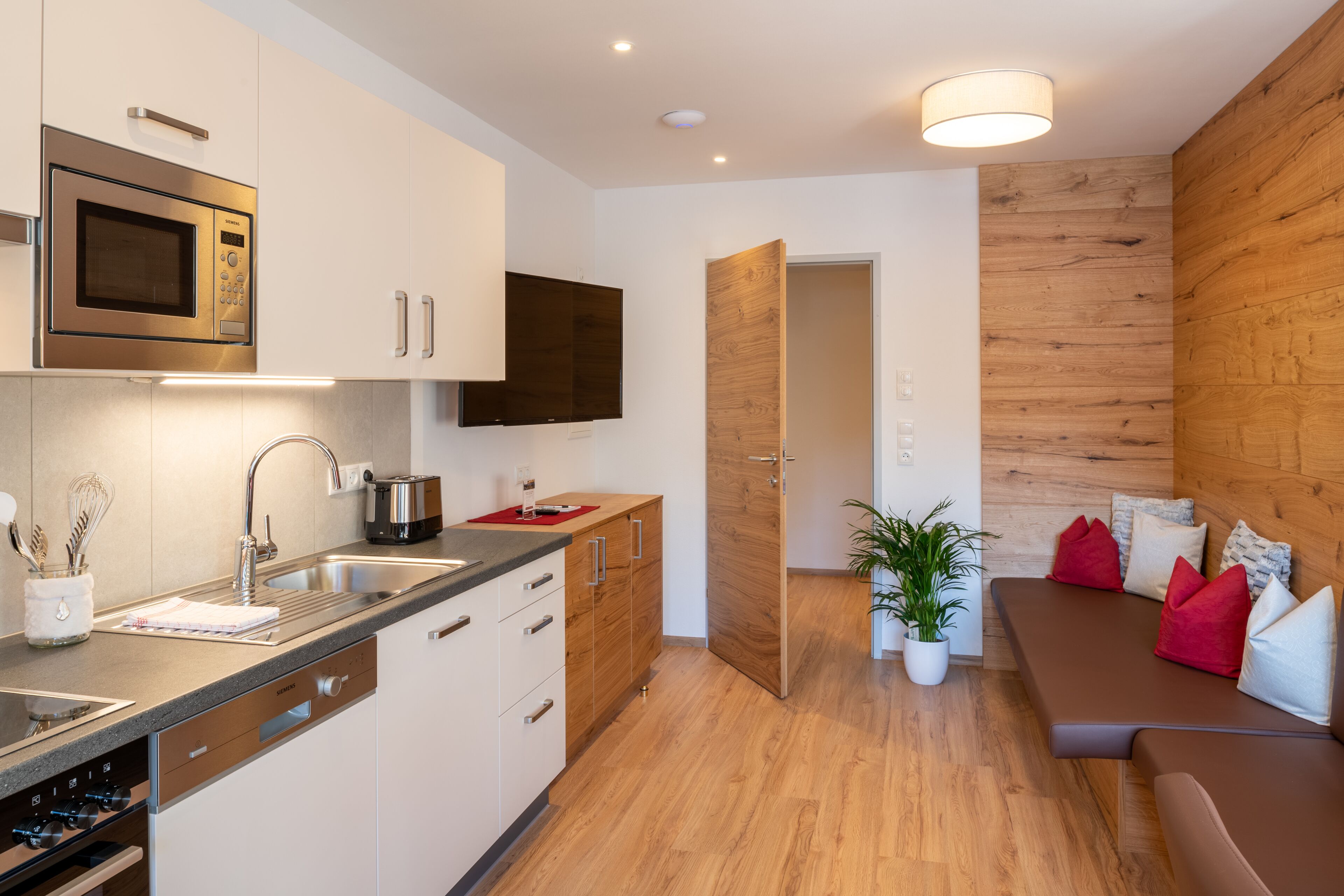 A modern kitchen with a built-in microwave, stainless steel sink, and wooden cabinets. There is a dining area with a brown sofa and red cushions.