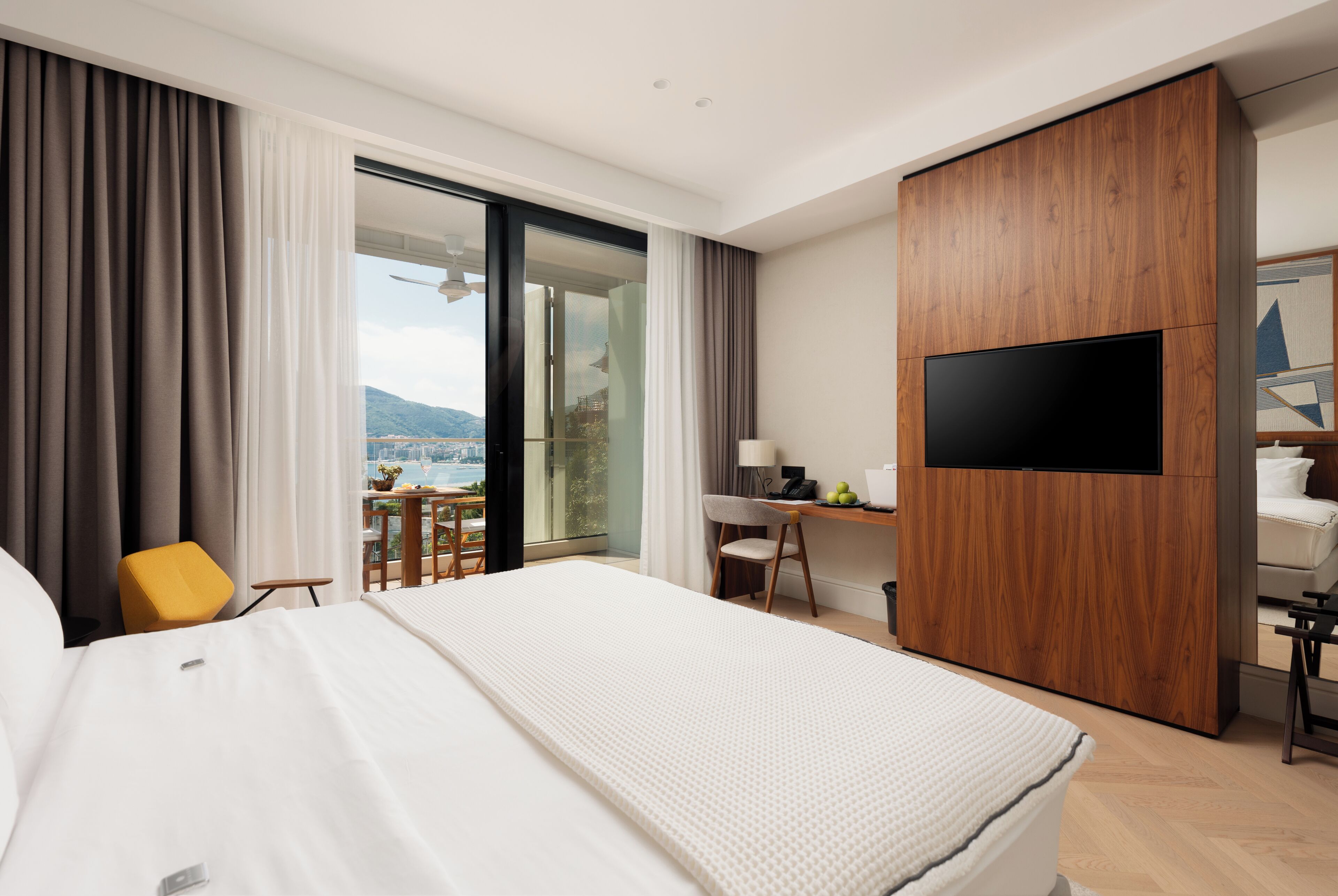 A hotel room with a large bed, a desk with a chair, a television, and a balcony with a view.