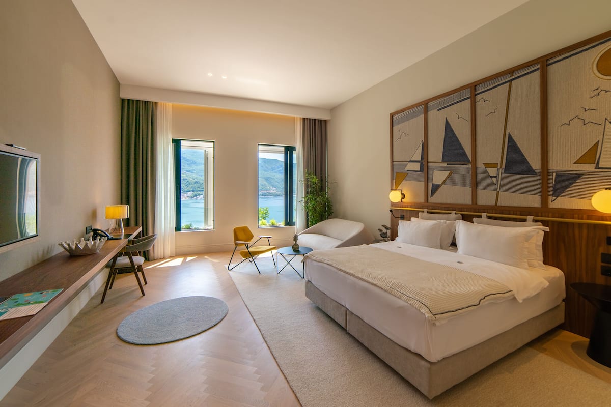 A hotel room with a large bed, a desk with a chair, a TV, and a view of the sea.