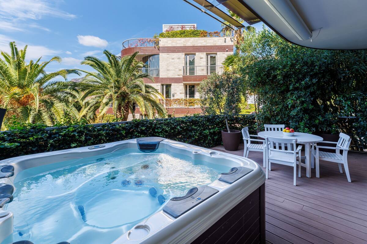 A hot tub on a wooden deck with a table and chairs, surrounded by greenery and a building in the background.