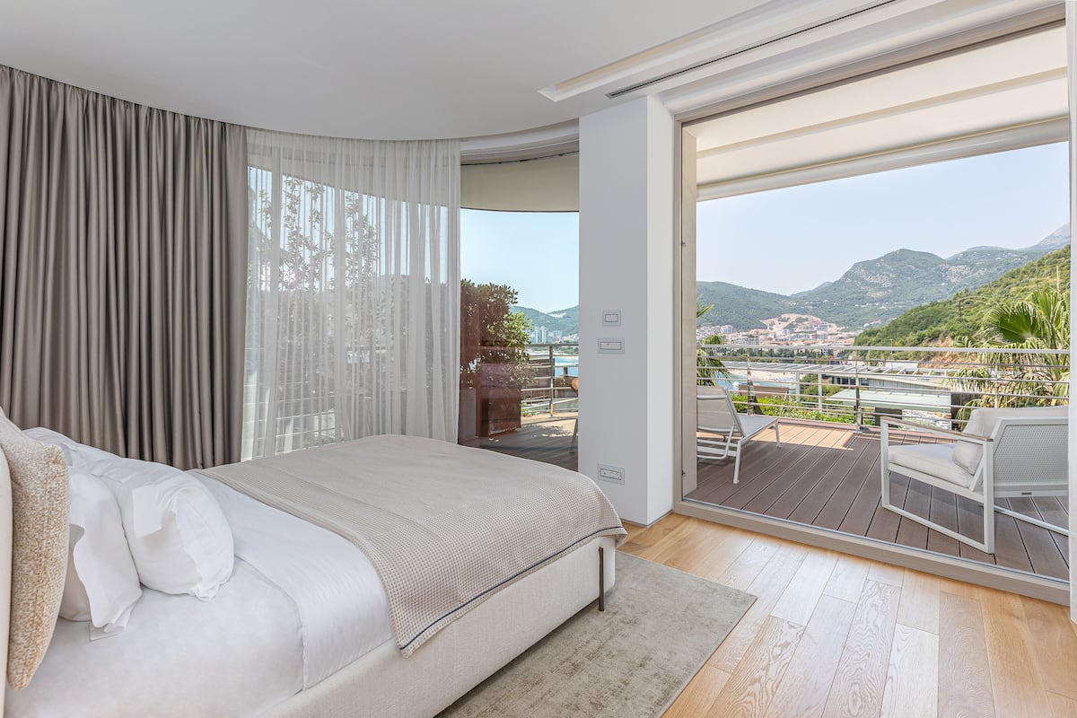 A bedroom with a large bed, a view of the balcony, and a cityscape in the background.