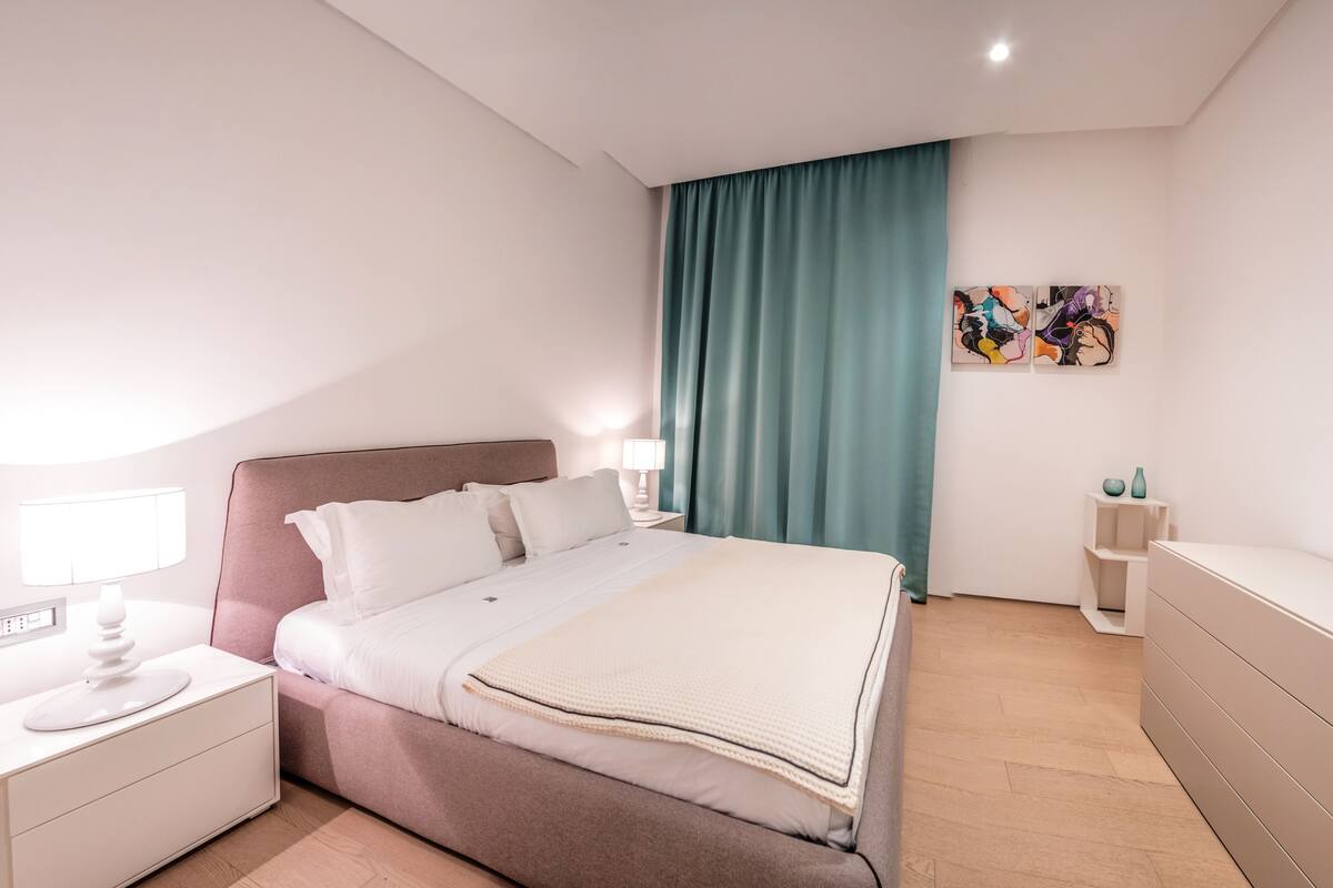 A modern bedroom with a bed, bedside tables, a nightstand, a dresser, and a window with curtains.