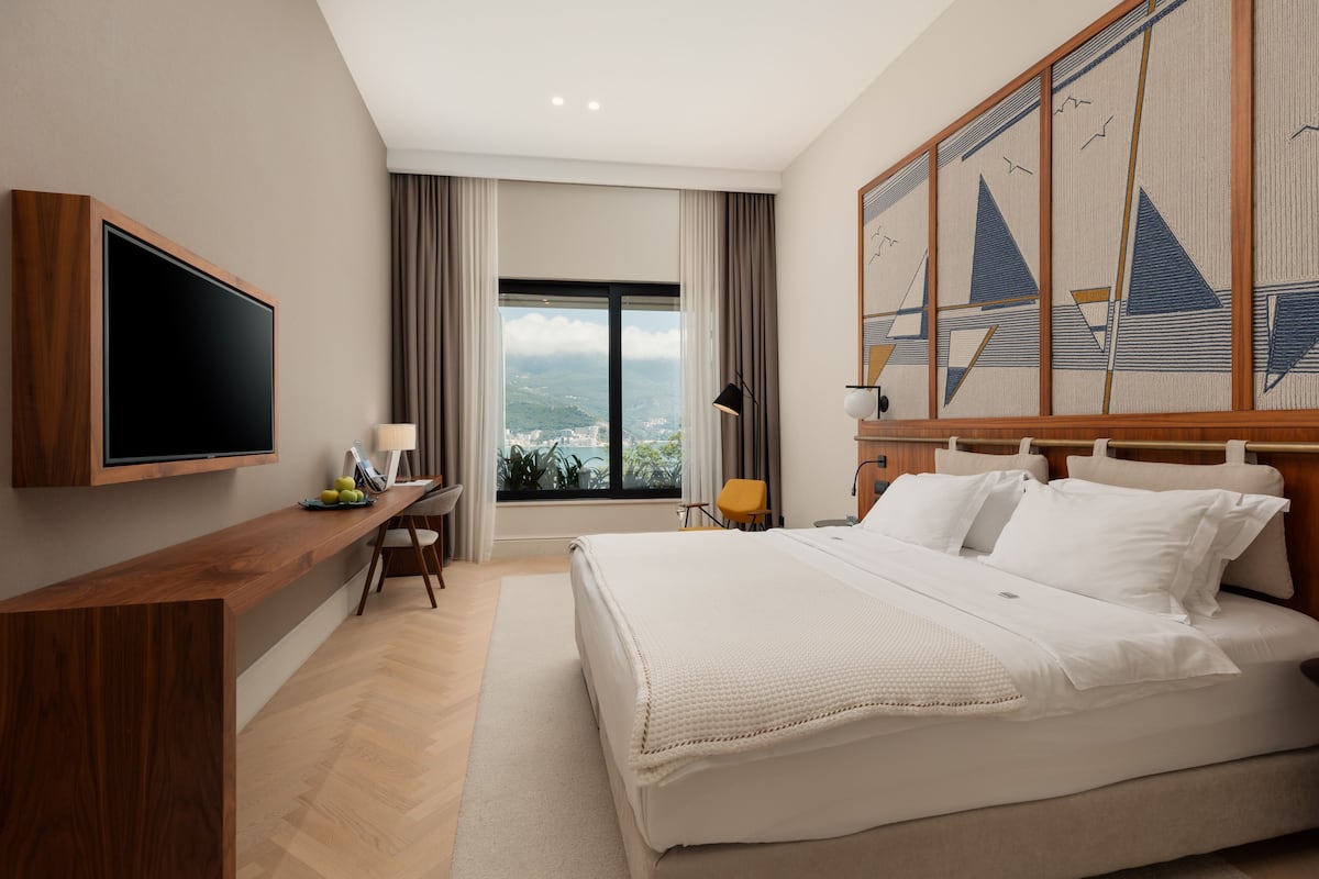 A modern hotel room with a large bed, a flat-screen TV mounted on the wall, a desk with a chair, and a large window offering a city view.