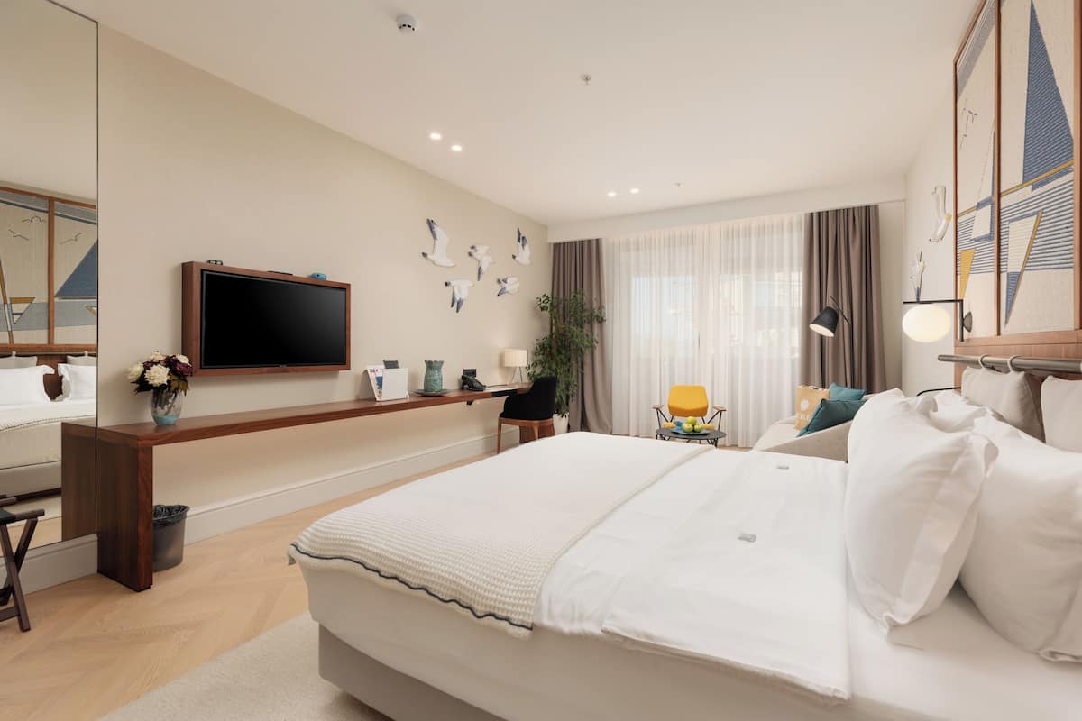 A modern hotel room with a large bed, a flat-screen TV mounted on the wall, a desk with a chair, a potted plant, and a window with curtains.