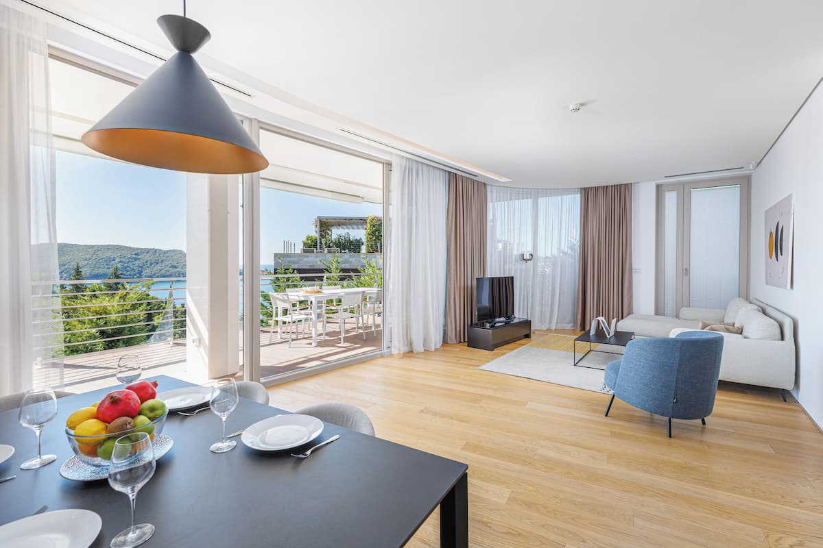 A modern living room with a sofa, a dining area with a table set for two, and a balcony with a view of the mountains.