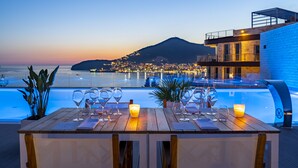 A dining table set with wine glasses and a candle, overlooking a pool and cityscape at dusk. - Dukley Hotel & Resort (Budva)