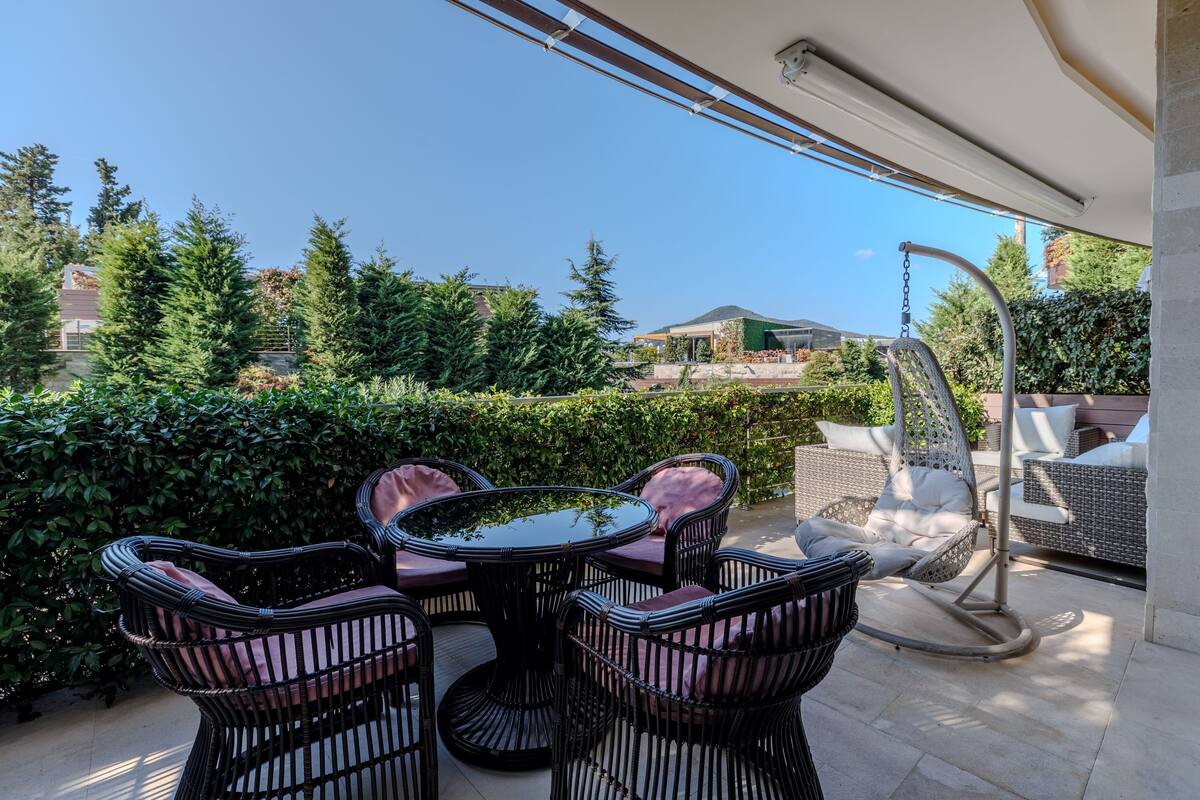 An outdoor patio with wicker furniture and a swing chair, surrounded by greenery and a clear sky.