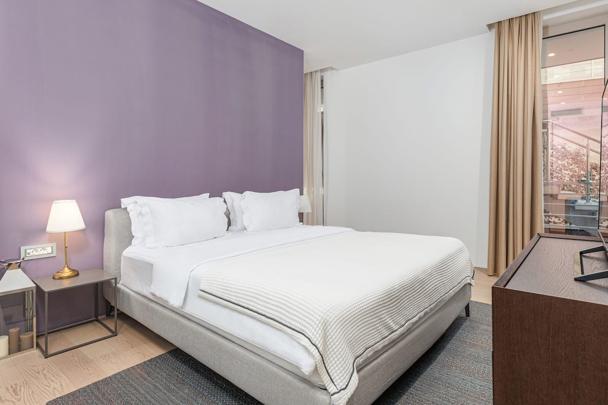 A hotel room with a bed, a nightstand with a lamp, a desk, and a view of the outside.