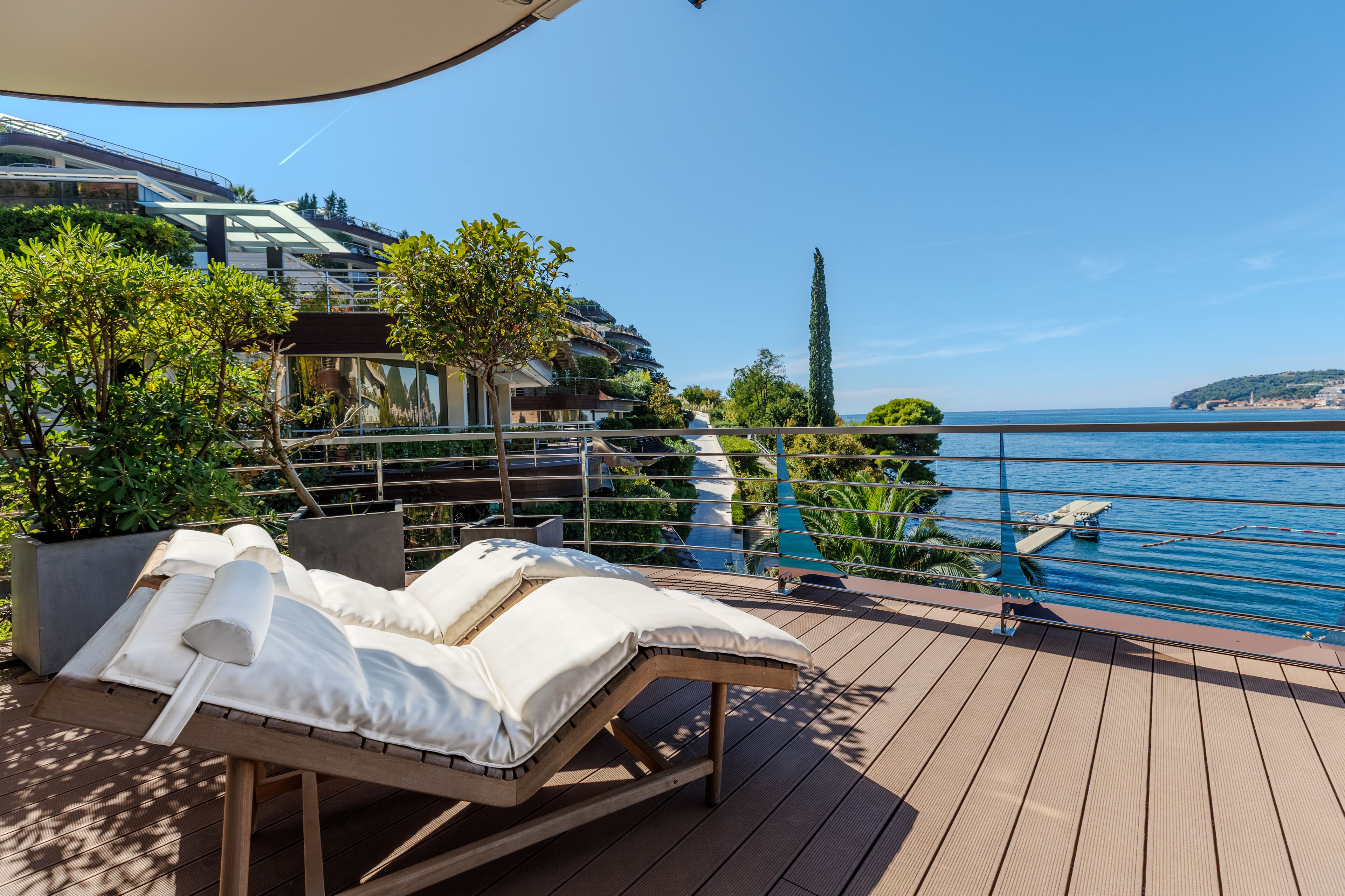 A sun lounger with white cushions on a wooden deck overlooking a pool and a coastal view.
