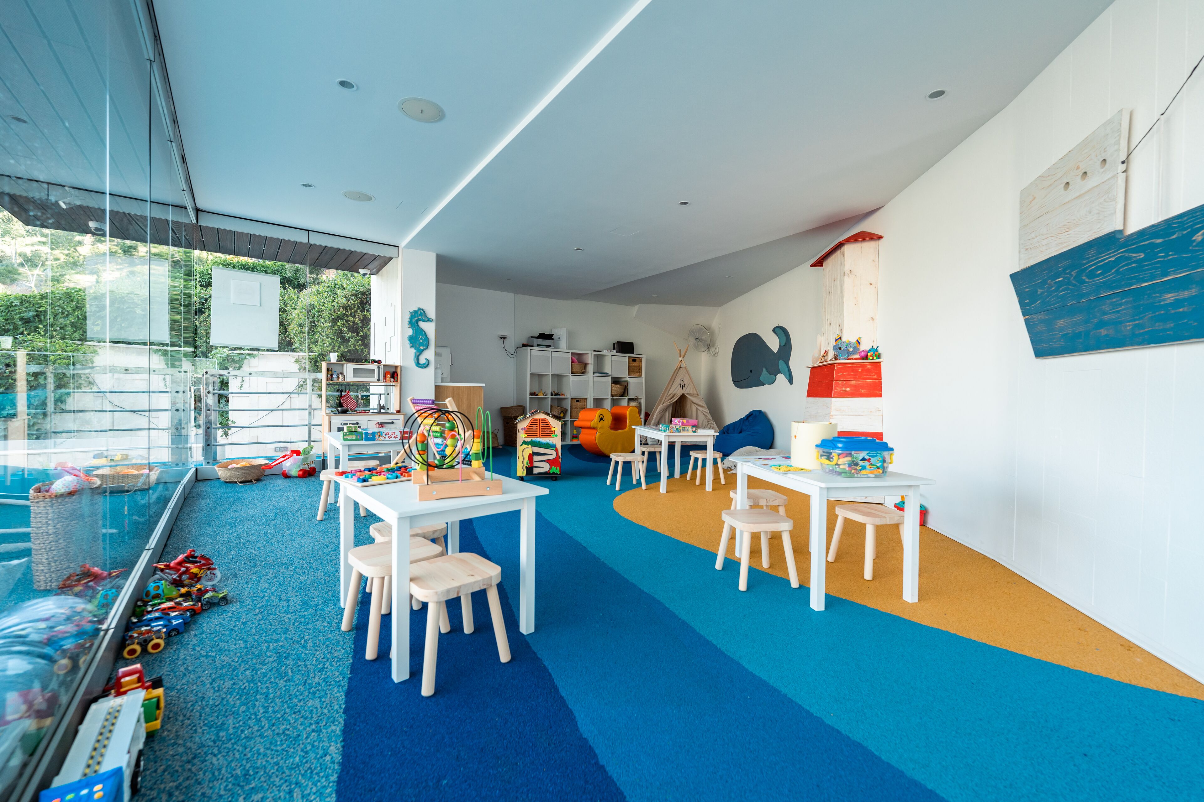 A children's playroom with a glass enclosure, play tables, chairs, and educational toys.