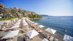 A beach with rows of sunbeds and umbrellas, a clear blue sea, and buildings in the background. - Dukley Hotel & Resort (Budva)