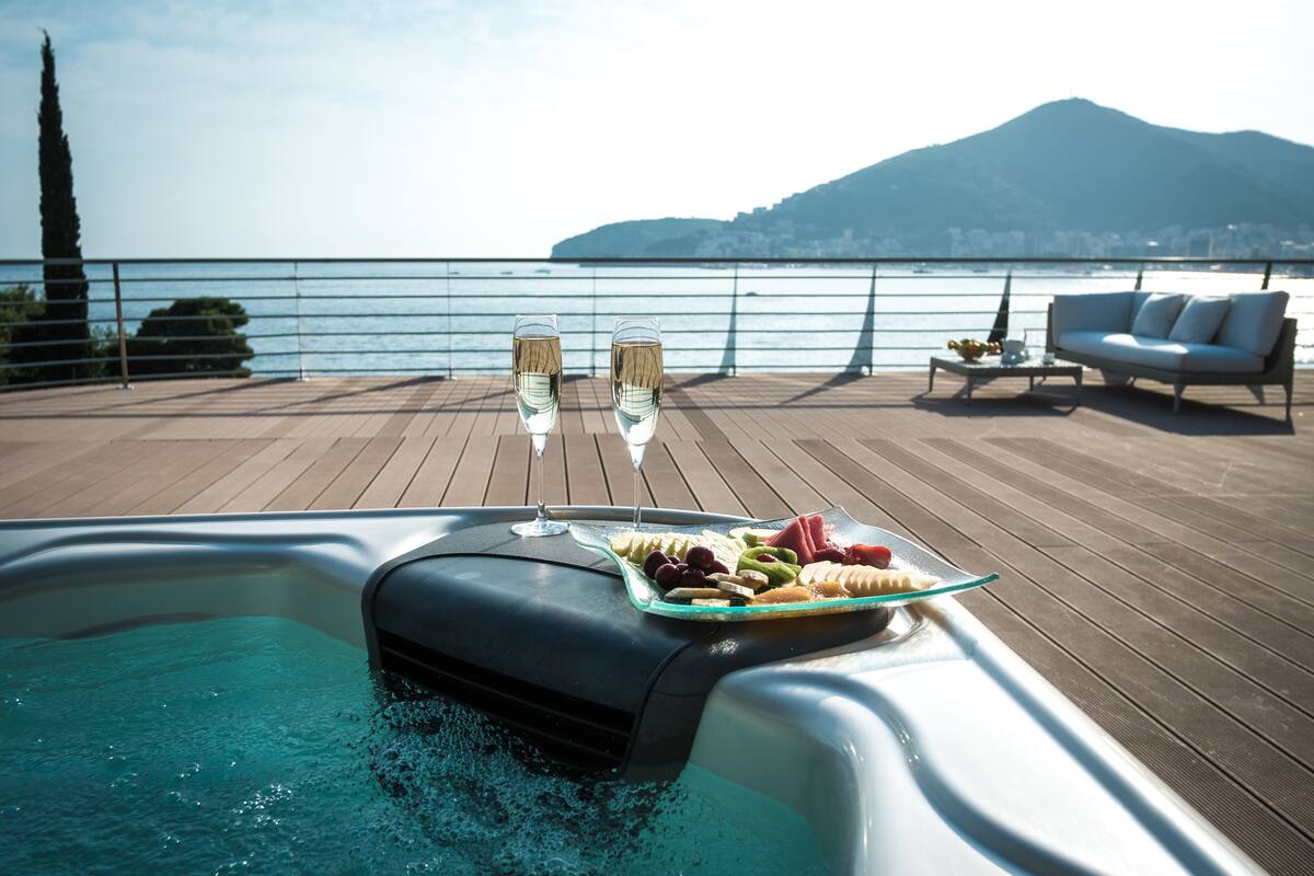 A hot tub with a glass tray of fruits and two glasses of wine, overlooking a body of water with a mountain in the background.