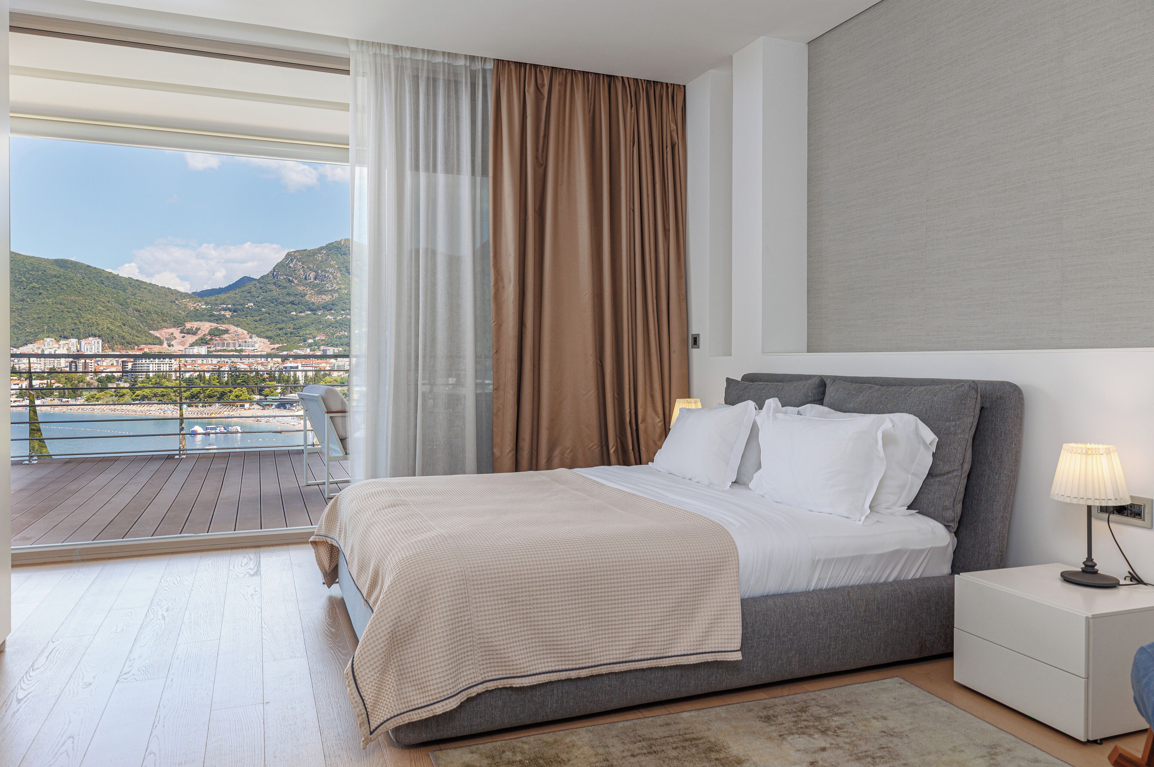 Une chambre avec un lit, une table de chevet, une lampe et une vue sur un balcon donnant sur une piscine et des montagnes.