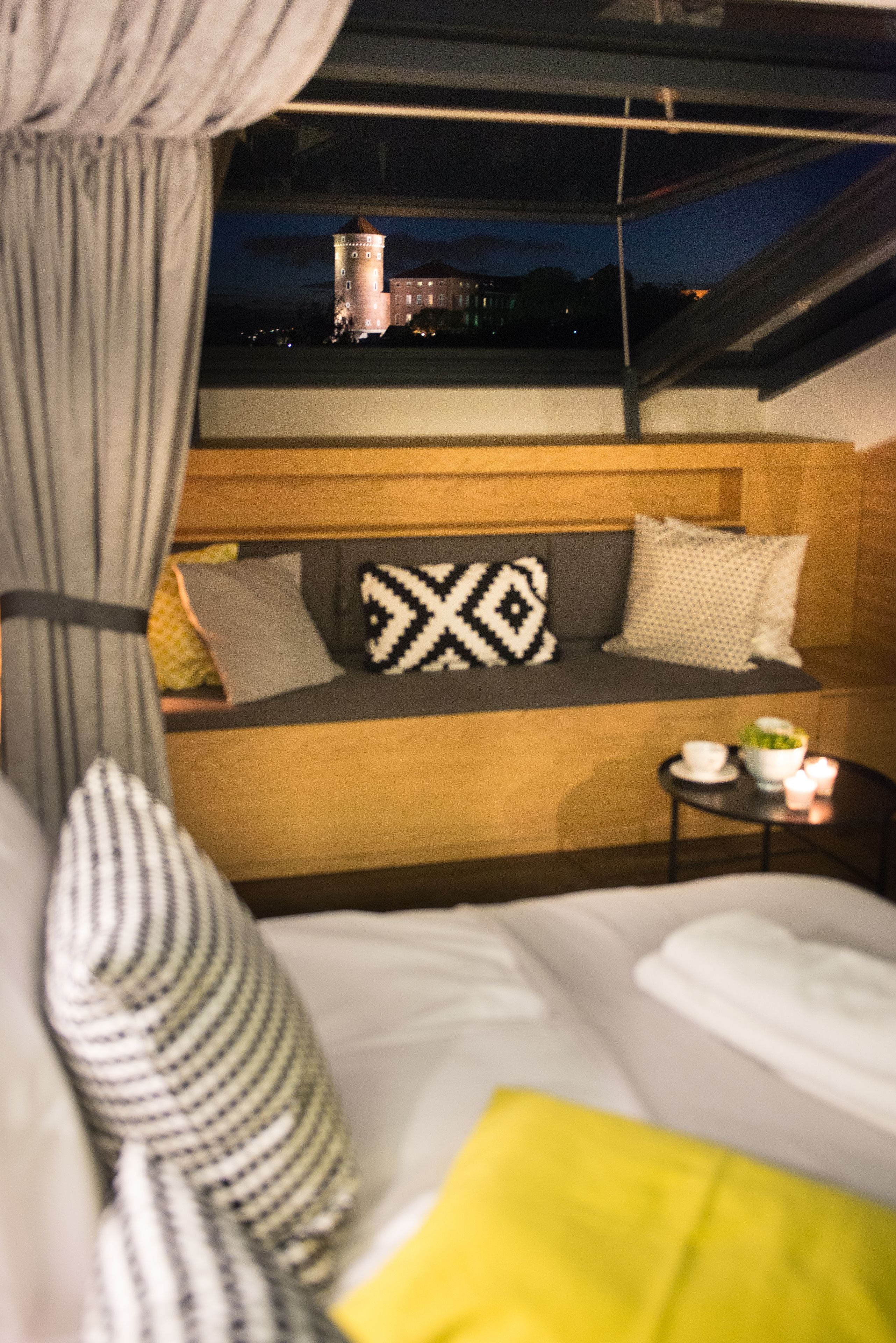Un dormitorio con cama, un banco de madera con cojines, una mesita con una vela y vista a una torre por la ventana.