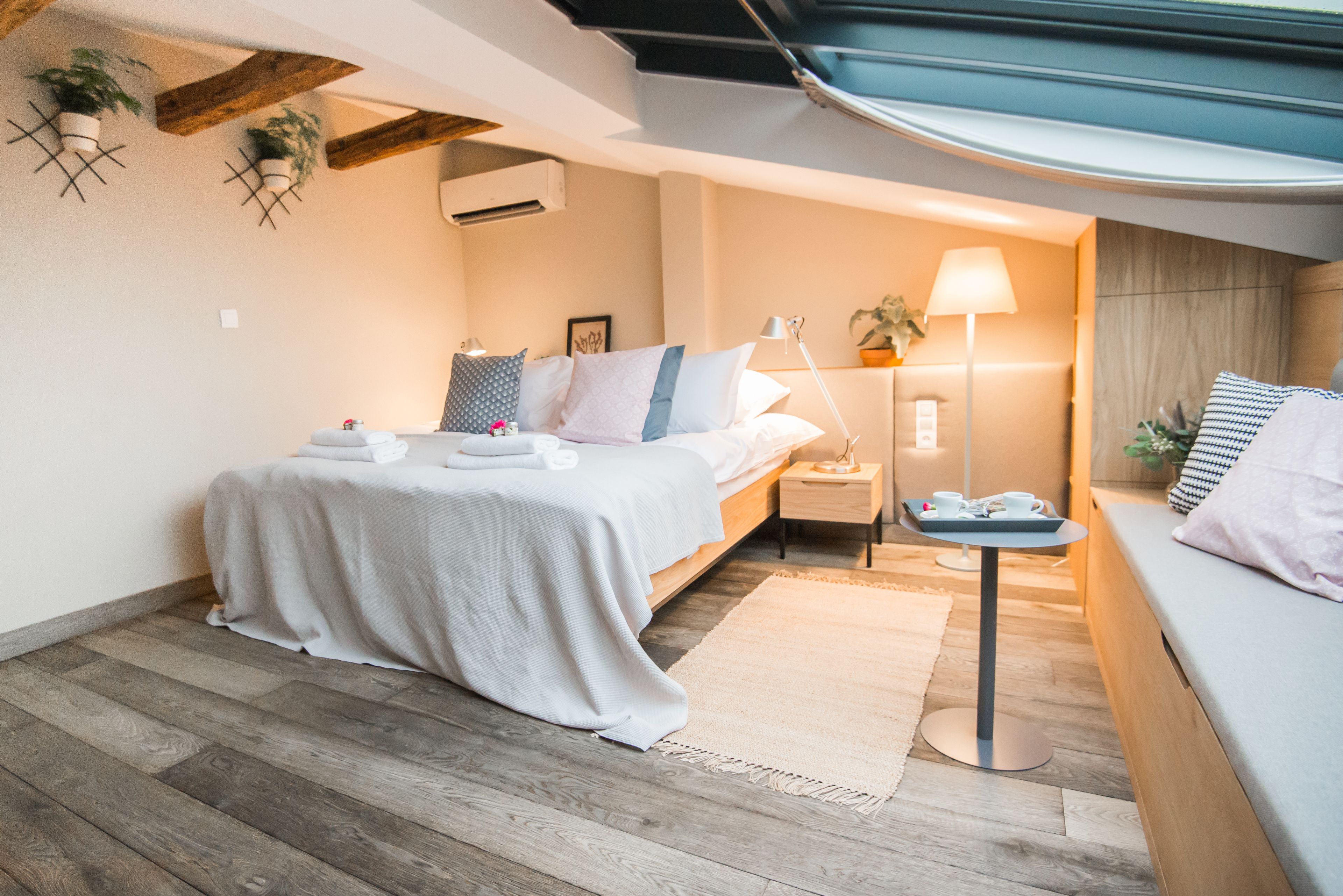 Quarto moderno com piso de madeira, cama com roupa de cama branca, mesa de cabeceira com abajur e uma pequena mesa com uma bandeja de objetos.