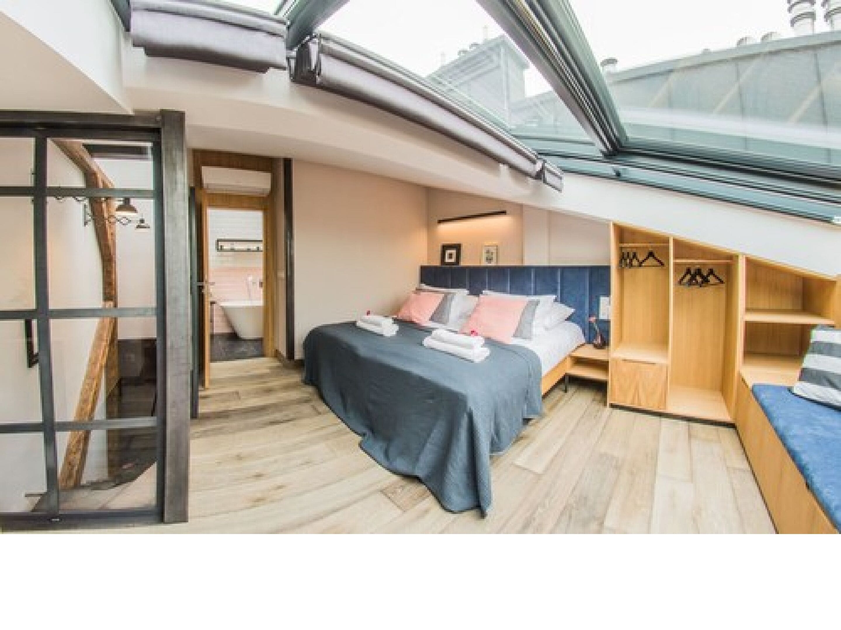 Ein kleines Schlafzimmer mit Oberlicht, einem Bett, einem Holzschrank und einem durch eine Glastür einsehbarer Badezimmer.