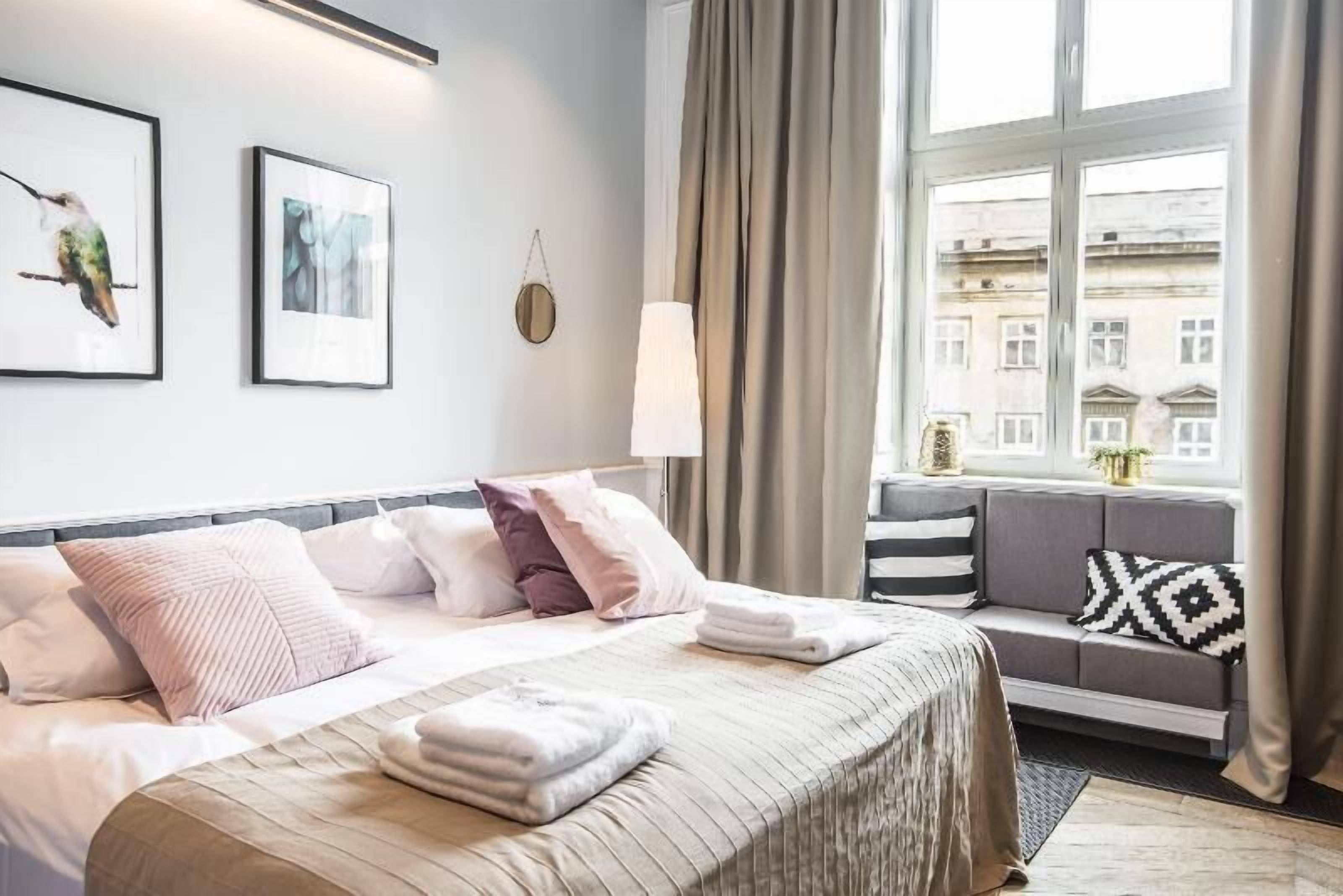 A hotel room with a bed, a sofa, a window with curtains, and artwork on the wall.