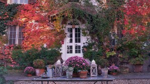 Un edificio con fachada blanca, ventanas de arco y una puerta. Hay plantas en macetas y una mesa con un farol y una regadera al frente.