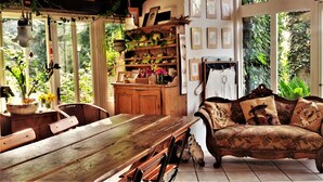 Una habitación acogedora con una mesa de comedor de madera, un sofá floral y una zona de cocina con estantes y plantas colgantes.